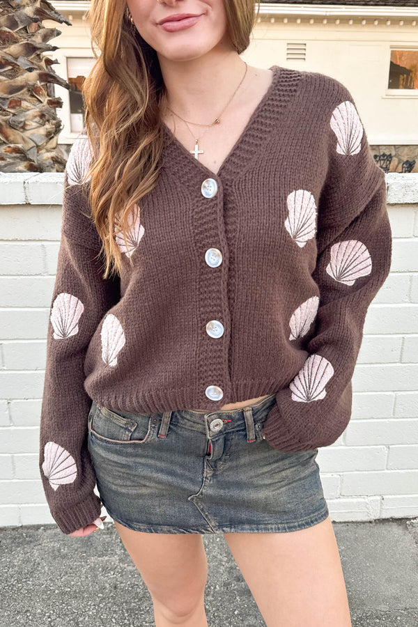 Woman wearing a brown cardigan with shell patterns in front of a white wall.